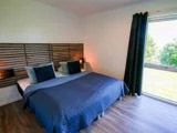 Et moderne soveværelse med dobbeltseng, blåt sengetøj, hovedgærde af træ, to sengelamper, mørke gardiner og et stort vindue med udsigt til grønne områder.