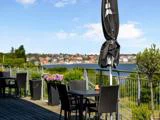Udendørs caféterrasse med sorte borde og stole, potteplanter og en lukket parasol med udsigt over et vandområde og en fjern by under en blå himmel.