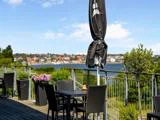 Udendørs terrasse med sorte kurveborde og -stole, potteplanter og en lukket parasol med udsigt over vand og en fjern by under en blå himmel.