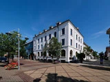 En hvid, treetagers historisk bygning med buede vinduer på et solrigt gadehjørne, omgivet af træer og blomsterbede.