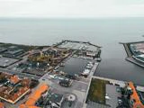 Luftfoto af en kysthavn med dokker, både og omkringliggende bygninger med orange hustage ved siden af det rolige hav under en overskyet himmel.