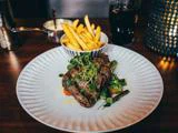 En tallerken med grillet bøf toppet med mikrogrønt, serveret med grøntsager, ved siden af en skål pommes frites og et glas mørk sodavand på et træbord.