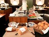 En morgenmadsbuffet med diverse brød, rundstykker, pålæg, en korndispenser og overdækkede bakker med skiveskåret frugt og kød på en disk i et veloplyst spiseområde.