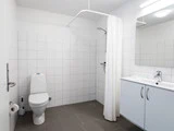 Minimalistisk badeværelse med hvide fliser, et toilet, en brusekabine med hvidt forhæng, et stort spejl og et hvidt vaskeskab.