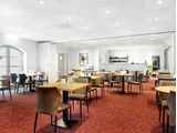 En moderne, veloplyst restaurant med træborde, brune stole, rødt gulvtæppe og et buffetområde bagved.