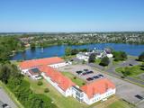 Luftfoto af bygninger med røde tage og en parkeringsplads nær en sø med en by og grønne områder i baggrunden under en klar blå himmel.