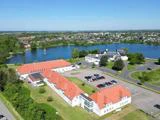 Luftfoto af bygninger med røde tage ved siden af en parkeringsplads, nær en sø med træer og en by i baggrunden under en klar blå himmel.