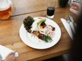 En hvid tallerken med en gourmetret med ris, grønt og skiveskårne grøntsager på et træbord med drikkevarer og redskaber i nærheden.