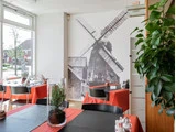 En hyggelig restaurant med røde duge, sorte stole og et stort vægmaleri af en vindmølle. På bordene står en plante og en glasdekoration, og sollyset strømmer ind gennem vinduet.
