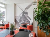 Et moderne restaurantinteriør med røde duge, sorte stole, en stor potteplante og et vægmaleri af en traditionel vindmølle.