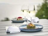 To elegante tallerkener med gourmetmad og to glas hvidvin er dækket på et udendørs bord med en sløret naturskøn baggrund.