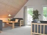 En hyggelig stue med skråt træloft, to sofaer, et rundt sofabord i træ med blomster, en høj plante og store vinduer, der lukker naturligt lys ind.