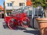 En række knaldrøde cykler med kurve er parkeret ved et metalrækværk på en solrig gade ved siden af en stor potteplante og bygninger i baggrunden.