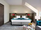 Moderne hotelværelse med dobbeltseng, grønt og hvidt sengetøj, to natborde med lamper, et lille bord med stole og et stort vindue, der lukker naturligt lys ind.