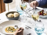 En person sidder ved et bord og spiser suppe af en hvid skål, med vinglas, en skål med brød og en tallerken med mad ved siden af.