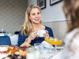 En kvinde med blondt hår sidder ved et bord og smiler, mens hun holder en hvid kaffekop i hånden.