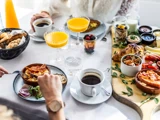En person spiser morgenmad ved et bord, der er dækket med skåle med æg og bacon, frugtkopper, croissanter og glas med appelsinjuice.