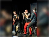 Tre musikere optræder på scenen: én synger i en mikrofon, én spiller elektrisk guitar, og én spiller bas. Alle er optaget af musikken i scenebelysningen.