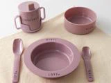 Et sæt lyserødt børneservice med skål, tallerken, kop med låg, gaffel og ske, hver især mærket med ord som MINI, LOVE og SWEETHEART, arrangeret på et beige klæde.