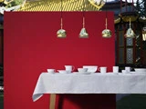 En hvid dug med hvide kopper og skåle er dækket foran en rød væg med fire guldpendler over og en traditionel asiatisk bygning i baggrunden.