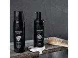 To sorte flasker med Meraki Mini badeprodukter og en hårbørste af træ står på en rustik hylde; skummet er synligt nær flaskerne mod en grå baggrund.