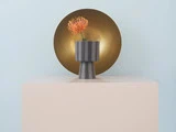 En enkelt orange blomst i en moderne, stribet grå vase står på en beige piedestal med en stor, rund guldskive i baggrunden mod en lyseblå væg.