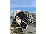 Et sort- og hvidstribet håndklæde er draperet over en træbænk ved en søstrand under en klar blå himmel.