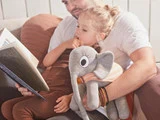 En mand læser en bog for en ung pige, der sidder på hans skød med en grå tøjelefant i hånden, mens de begge slapper af i en sofa.