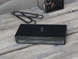 En sort, rektangulær glaskasse med låg på en forvitret træoverflade, ved siden af to store sorte papirclips og en dekorativ lanterne.