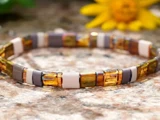 Et perlearmbånd med skiftevis firkantede perler i ravfarvede, brune og hvide toner på en stenoverflade med en gul blomst i baggrunden.