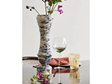 En høj, marmoreret keramikvase med lyserøde blomster står på et reflekterende bord ved siden af et vinglas, et mønstret glas og en foldet rustfarvet serviet.