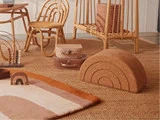 Et hyggeligt legeværelse med kurvestol, tremmeseng, vintagekufferter, regnbueformet pude, kugleramme og et stribet tæppe på en vævet måtte.