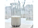 En klar glaskande med en lille mængde mælk står på en lys træoverflade med en sløret udendørs baggrund.