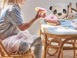Et lille barn sidder ved et lille bord med en lyserød donut i hånden, en anden donut på en tallerken og udstoppede panda- og hundelegetøj på bordet.