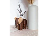 En lille elefantfigur i træ med lyse og mørke trætoner står på en lys træoverflade ved siden af en høj, mat lysegrå vase. Tørrede grene ses i baggrunden.