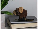 En lille elefantfigur i træ sidder på en stak bøger og notesbøger med en kuglepen ovenpå. En grøn plante er synlig på venstre side.