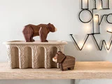 To geometriske bjørnefigurer af træ på en træhylde; den ene står på en tekstureret beholder, den anden ved siden af. I baggrunden ses et dekorativt vægstykke med lyskæder.