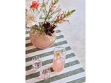 En vase med forskellige blomster og en kande lyserød limonade med to glas står på en grøn- og hvidstribet dug.