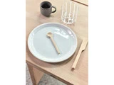 En hvid tallerken med en lille træske, en trækniv ved siden af, et klart glas og en grå kop på et lyst træbord.