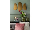 En moderne stue med grønne vægge, abstrakte guldspejle, en vase med blomster, stablede bøger, en fuglefigur og strukturerede rosa og cremefarvede puder på en grå sofa.
