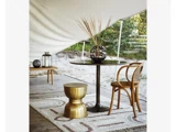 Et lille rundt bord med en dekorativ vase, en gylden skammel, en træstol og et mønstret tæppe på en træterrasse under en hvid baldakin. I baggrunden ses udendørs grønne områder.