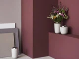 Tre vaser, den ene med farverige vilde blomster, står på geometriske bordeauxrøde og beige overflader mod en lilla væg med stablede paneler i baggrunden.