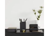 Et minimalistisk sort skrivebordsorganizer-sæt med kuglepenne, en bakke, æsker og guldclips står på en sort overflade; en hvid vase med grønne blade står i baggrunden.