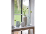 Hvide og blå glasvaser med sarte blomster og et hvidt muslingeskalsornament står i en vindueskarm med gennemsigtige gardiner og udsigt til et grønt udendørslandskab.