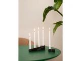 Fem tændte hvide stearinlys i sorte marmorholdere står på et grønt bord med en bladplante og en beige stol i baggrunden.