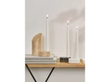 Tre tændte hvide stearinlys i minimalistiske stager står på en træhylde sammen med stablede bøger og et dekorativt stenstykke mod en almindelig lys væg.