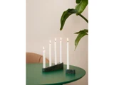 Fem tændte hvide stearinlys i marmorholdere er placeret på et grønt bord ved siden af en stor bladplante og en beige stol mod en lys væg.