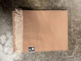 Et foldet tæppe af beige uld med cremefarvede frynser og et lille, påsyet mærke.