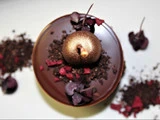 En blank chokoladedessert toppet med en guldfarvet kugle, rød og lilla pynt og chokoladekrummer med spredte dekorationer på en hvid overflade.