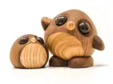 To uglefigurer af træ med store runde sorte øjne, en stående og en liggende på siden, på en hvid baggrund.